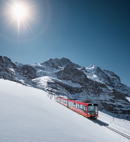 Jungfraubahn Winter | © Jungfraubahnen, Jeroen Seyffer Fotografie Ein roter Zug fährt eine verschneite Berghangstrecke entlang, umgeben von majestätischen, schneebedeckten Gipfeln unter klarem, blauem Himmel. Die Sonne scheint hell und taucht die Winterlandschaft in warmes Licht. | © Jungfraubahnen, Jeroen Seyffer Fotografie