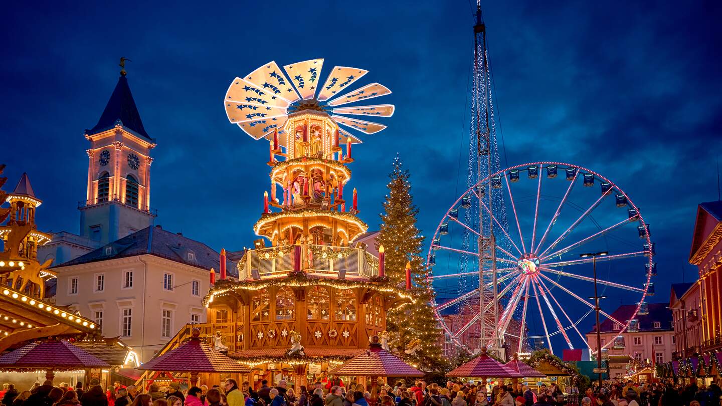 Festliche Szene auf einem Weihnachtsmarkt mit einer großen, beleuchteten Weihnachtspyramide, einem hell erleuchteten Riesenrad im Hintergrund und einem Kirchturm mit Uhr. Zahlreiche Besucher schlendern durch die abendliche Marktstimmung. | © KTG Karlsruhe Tourismus GmbH, Bruno Kelzer