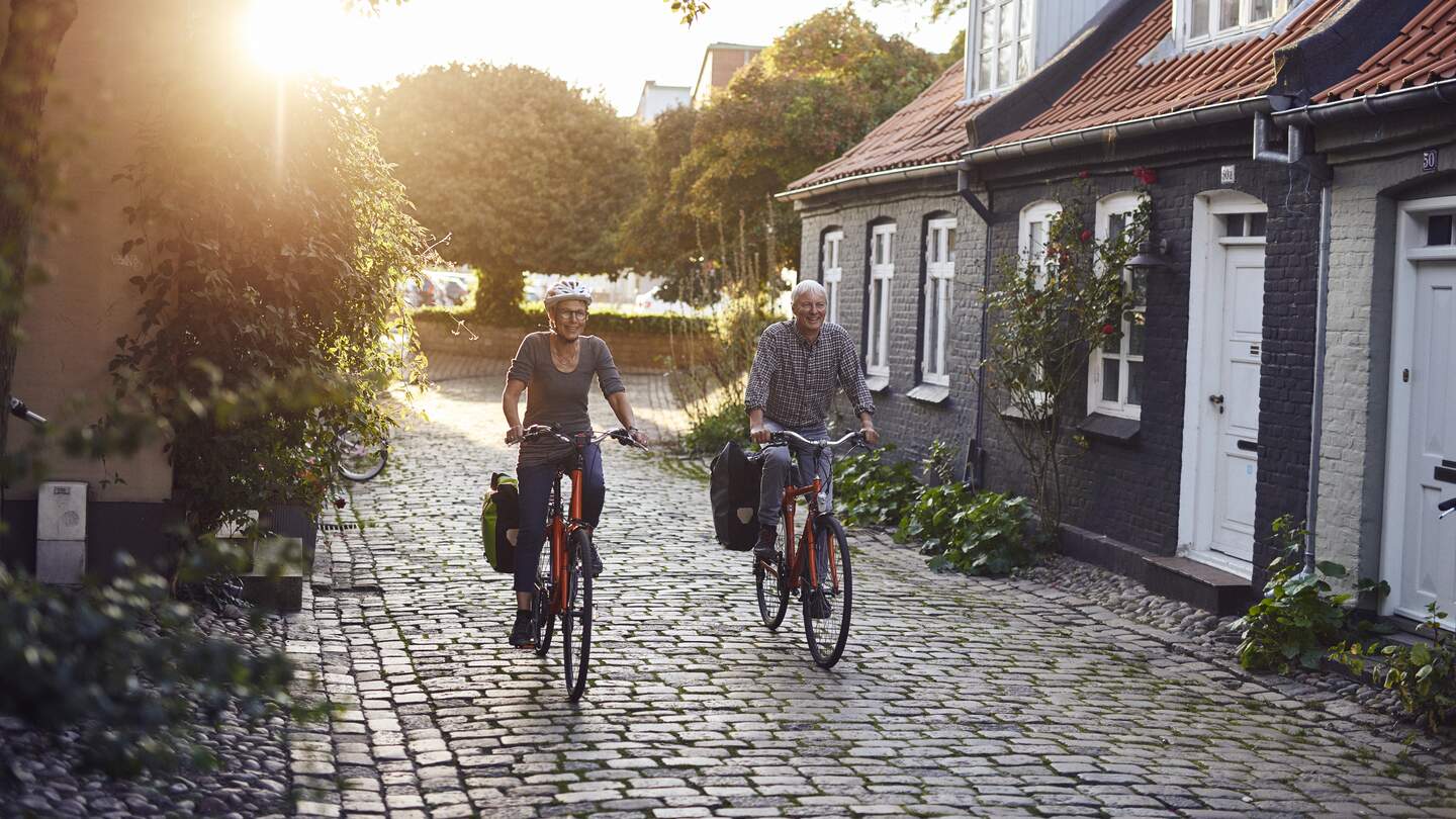 Zwei Radfahrende fahren durch eine sonnige Kopfsteinpflastergasse mit historischen Häusern in Aarhus. | © Niclas Jessen