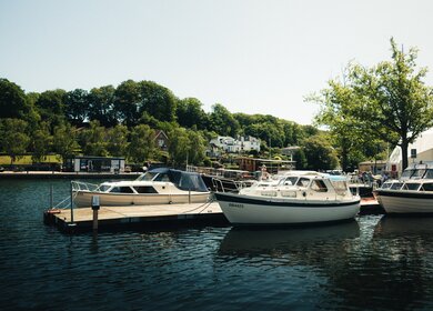 Boote liegen im Hafen von Silkeborg vor grüner Uferlandschaft und Wohnhäusern. | © Nikolaj Smed