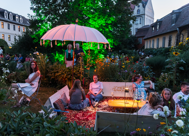 Ein kleiner Platz, umgeben von Häusern und begrühnt mit Bäumen und Pflanzen. Personen sitzen auf Bänken und stehen auf dem Platz. In der Bildmitte ist ein pinker Sonnenschirm. Es wird Goethes Geburtstag beim Weimarer Sommer gefeiert. | © Axel Clemens, weimar GmbH