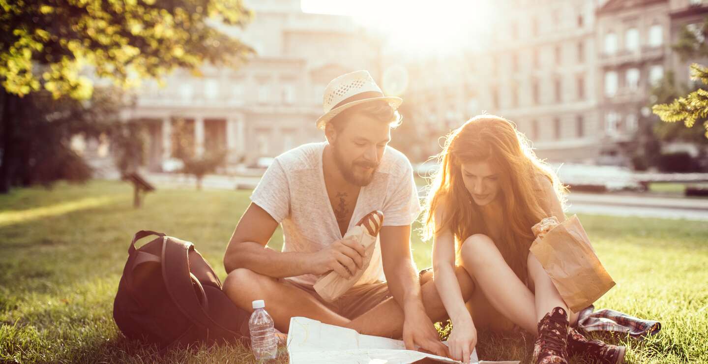Junges Touristenpaar, das die fremde Stadt erkundet. Auf dem Gras im Park sitzend, frühstückend und Stadtplan lesend. Tragen Freizeitkleidung. | © © Gettyimages.com/svetikd