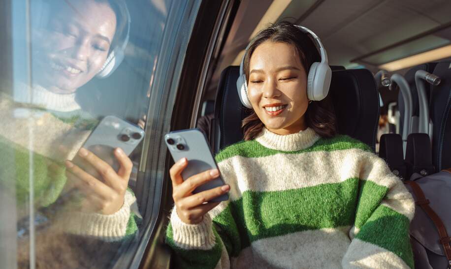 Person sitzt im Zug am Fenster, trägt weiße Kopfhörer und hält ein Smartphone in der Hand, mit Spiegelung im Glas und Tageslicht im Hintergrund. | © Gettyimages.com/	Milko