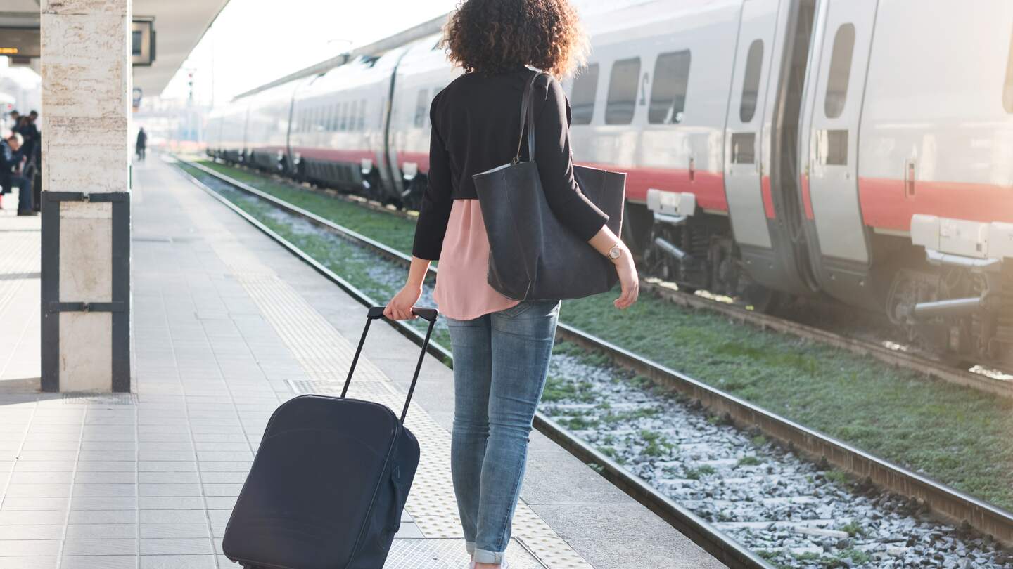Eine Frau mit dunklen Locken steht mit ihrem schwarzen Koffer am Bahngleis | © Gettyimages.com/tommaso79