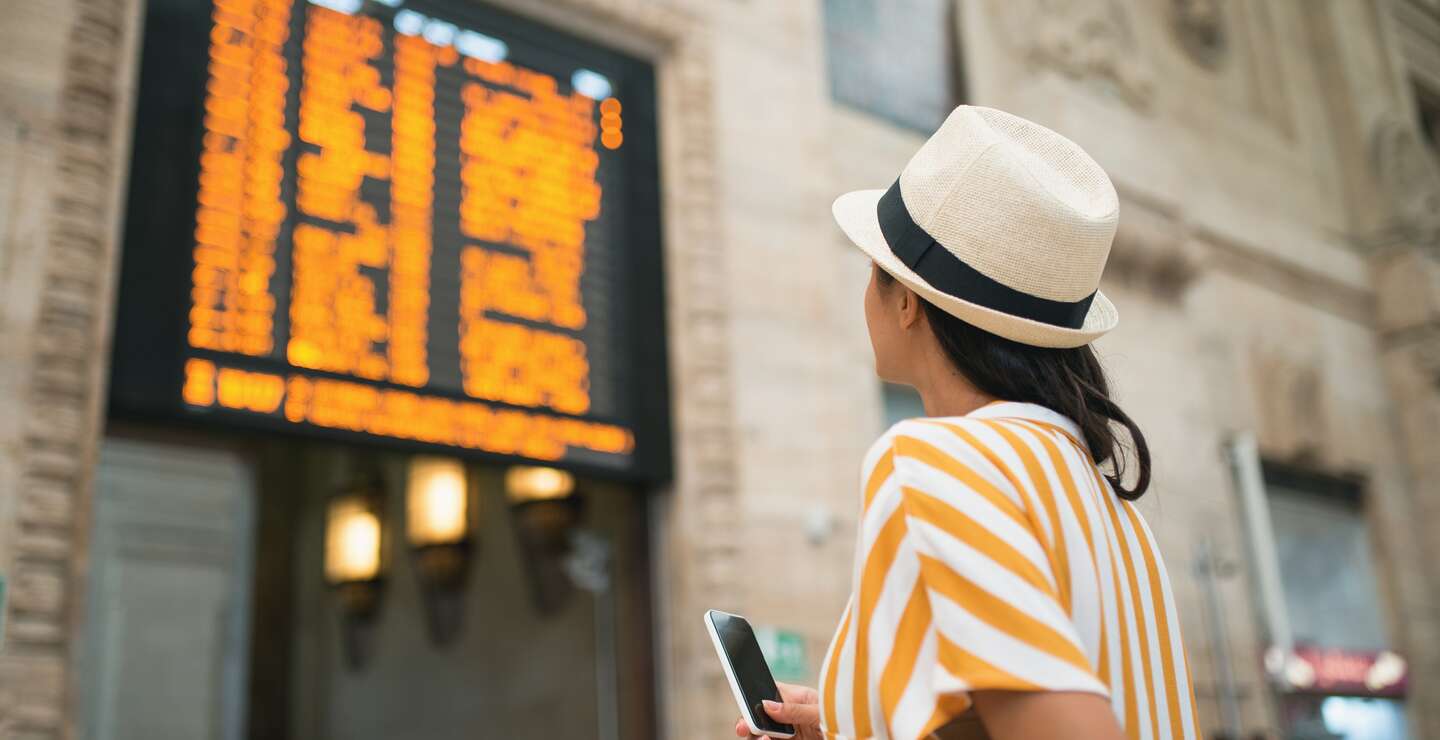 Eine dunkelhaarige Frau steht mit Sommerhut und Sommeroutfit an der Anzeigetafel der Bahn | © Gettyimages.com/MStudioImages