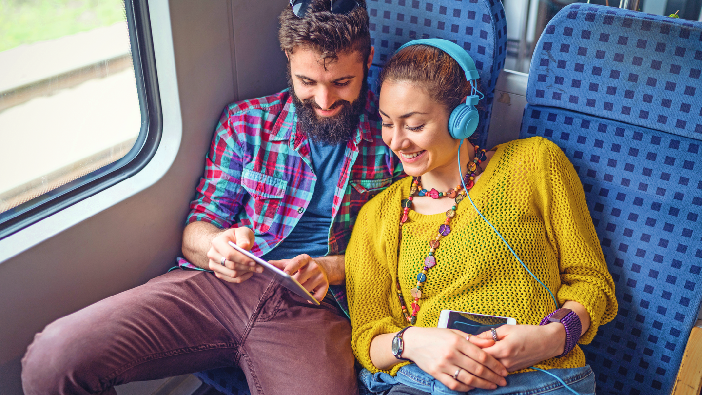Ein Pärchen sitzt mit Kopfhörern lachend im Zug und schauen sich etwas an | © Gettyimages.com/Mixmike