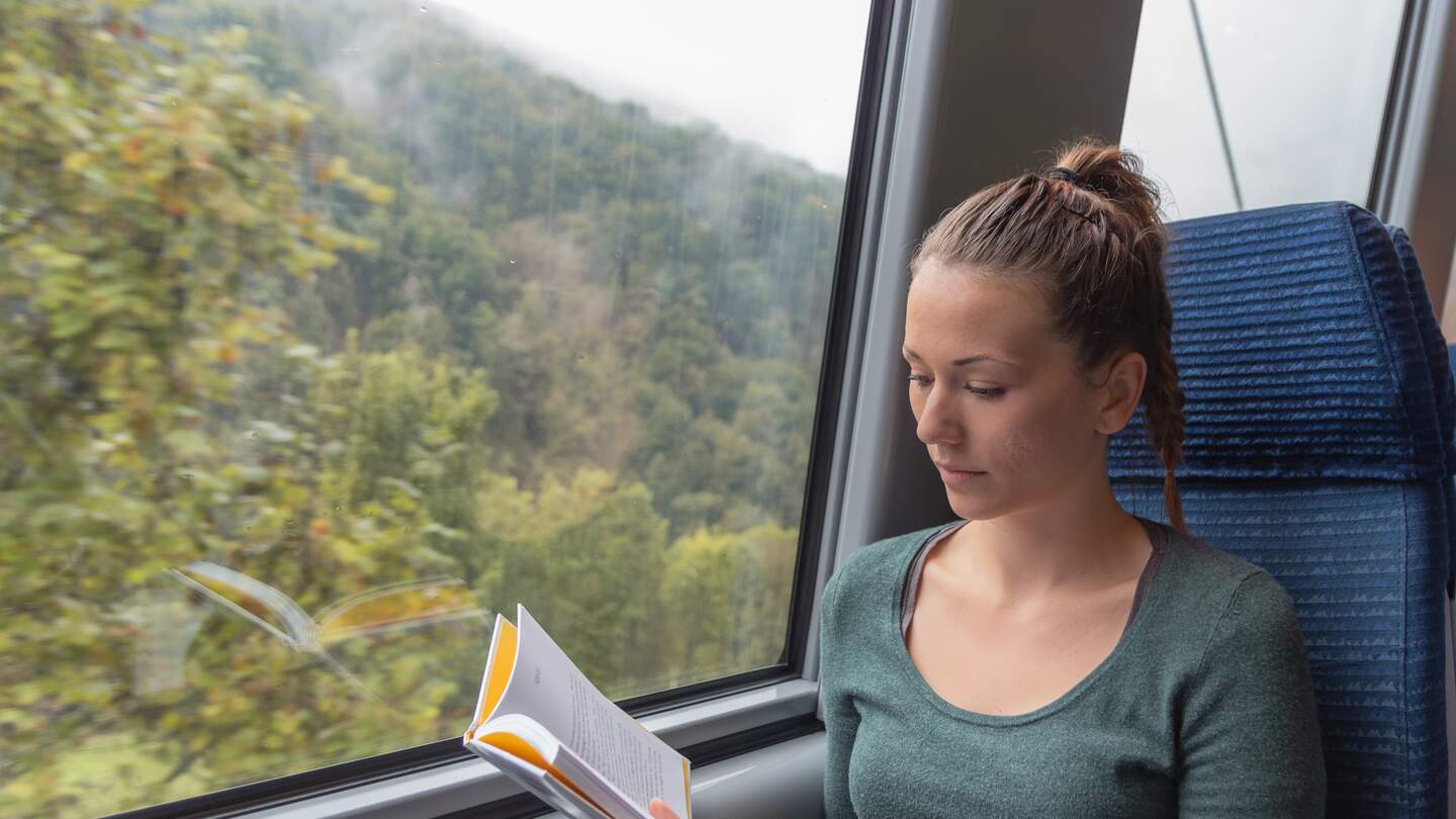 Eine blonde Frau mit geflochtenem Haar liest ein gelbes Buch im Zug | © Gettyimages.com/NDStock