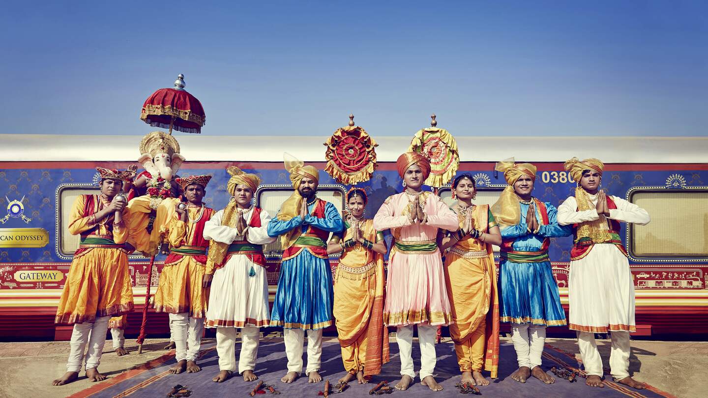 Eine Gruppe des Zugteams in farbiger, tradioneller indischer Kleidung, vor dem indischen Luxuszug Deccan Odyssey stehend | © Deccan Odyssey