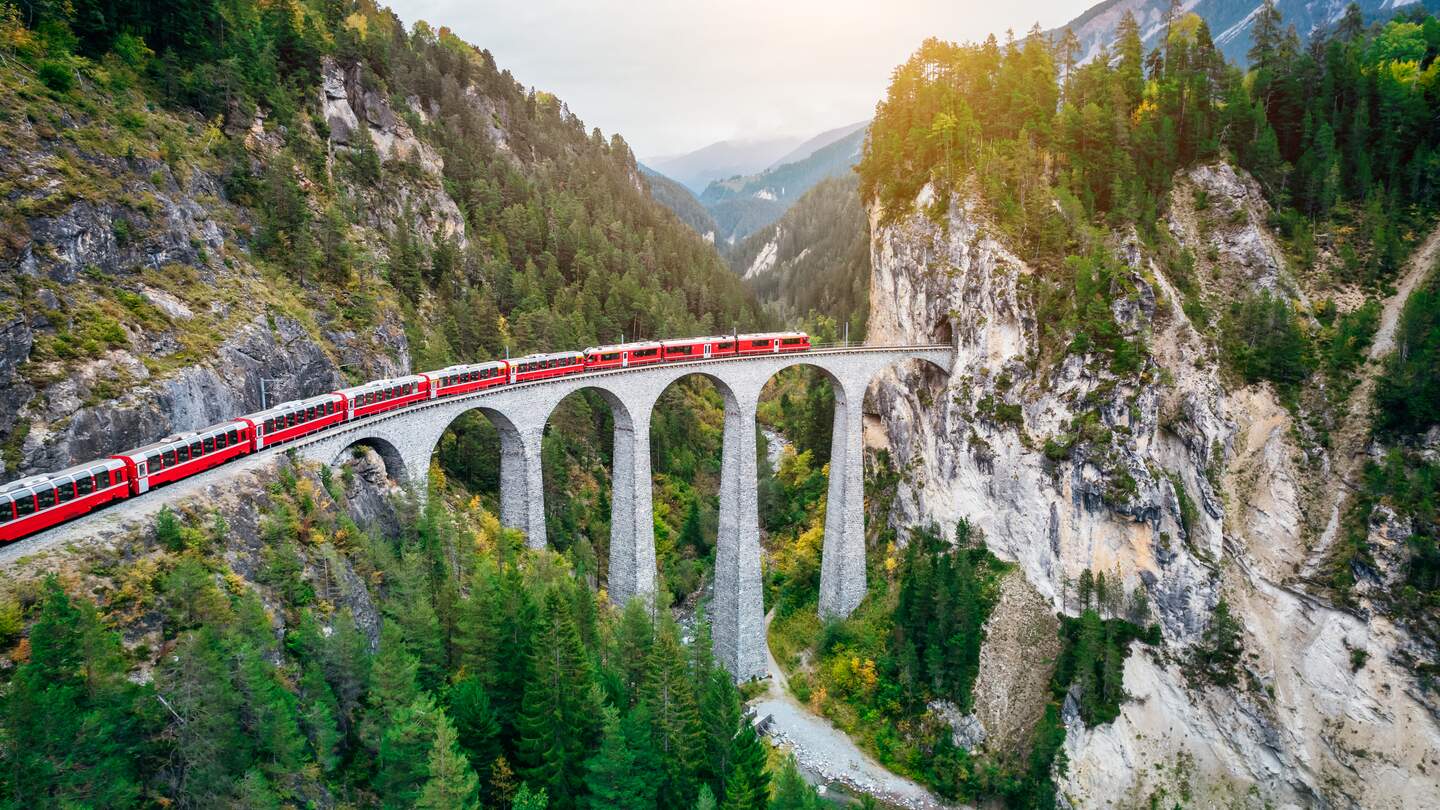 Bernina Express fährt über ein hohes Steinviadukt zwischen bewaldeten Felsen und Bergen in einer grünen Gebirgslandschaft. | © GettyImages.com/Nikada