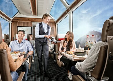 Zwei Paare sitzten im Speisewagen 1. Klasse mit Panoramafenstern und eine Mitarbeiterin bringt Wein | © Glacier Express AG/Stefan Schlimpf