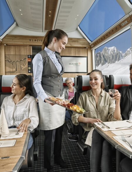 Zwei Paare sitzten im Speisewagen 2. Klasse mit Panoramafenstern und eine Mitarbeiterin bringt essen | © Glacier Express AG/Stefan Schlimpf