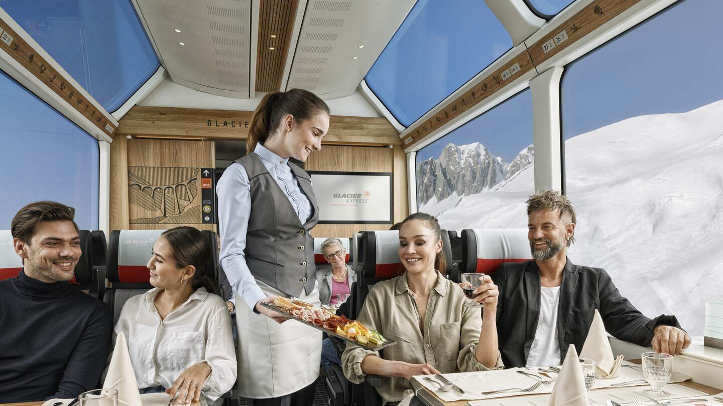 Zwei Paare sitzten im Speisewagen 2. Klasse mit Panoramafenstern und eine Mitarbeiterin bringt essen | © Glacier Express AG/Stefan Schlimpf