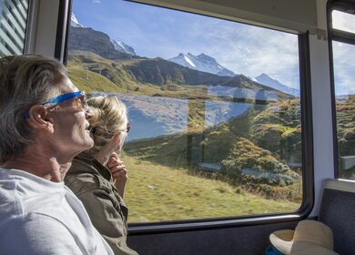 Bestager-Paar genießt den Blick aus einem Zugfenster, auf das sonnige Panorama der Schweizer Berge | © Gettyimages/AscentXmedia