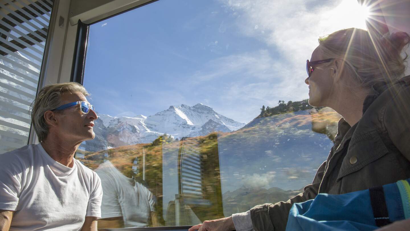 Bestager-Paar genießt den Blick, aus einem Zugfenster, auf das sonnige Panorama der verschneiten Schweizer Bergegipfel | © © Gettyimages/AscentXmedia
