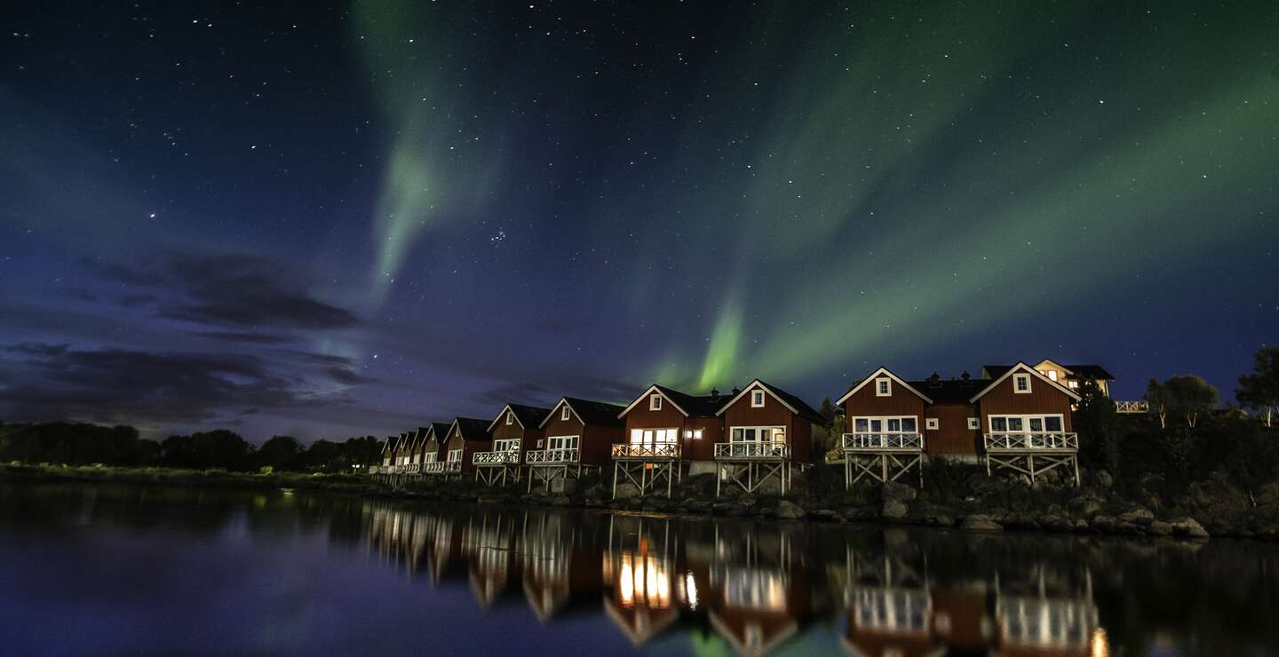Polarlichter, reflektiert im ruhigen Gewässer, über den roten Hütten in Nordnorwegen | © Gettyimages.com/Glenn Pettersen