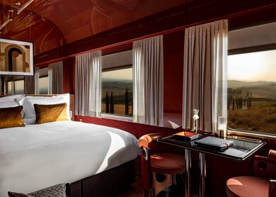 Blieck in die Suite des La Dolce Vita Orient Express mit großen Panoramafenstern, einem Doppelbett auf der linken Seite, und einem Tisch mit zwei Stühlen in der Mitte | © Orient Express SAS