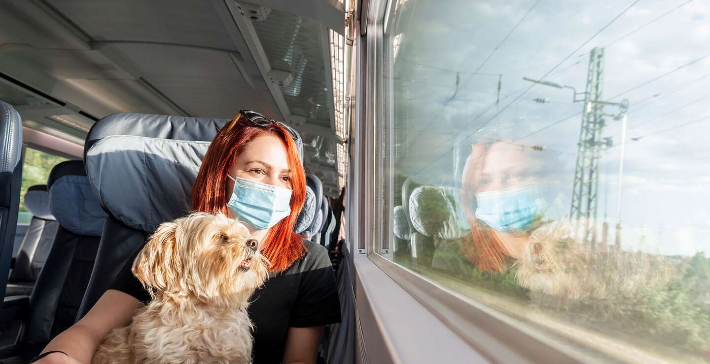 Rothaarige Reisende mit FFP2-Maske und kleinem Hund genießt die Aussicht vom Fensterplatz während der Bahnreise | © Gettyimages.com/YesPhotographers