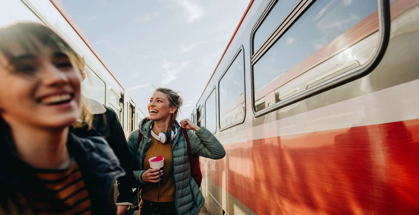 Zwei gut gelaunte, junge Frauen auf dem Bahnsteig zwischen zwei Zügen | © Gettyimages.com/AleksandarNakic