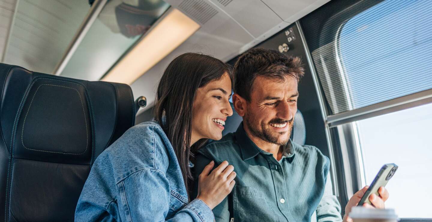 Pärchen im Zug an einem Tisch schauen auf ein Handy | © GettyImages.com/miloradkravic