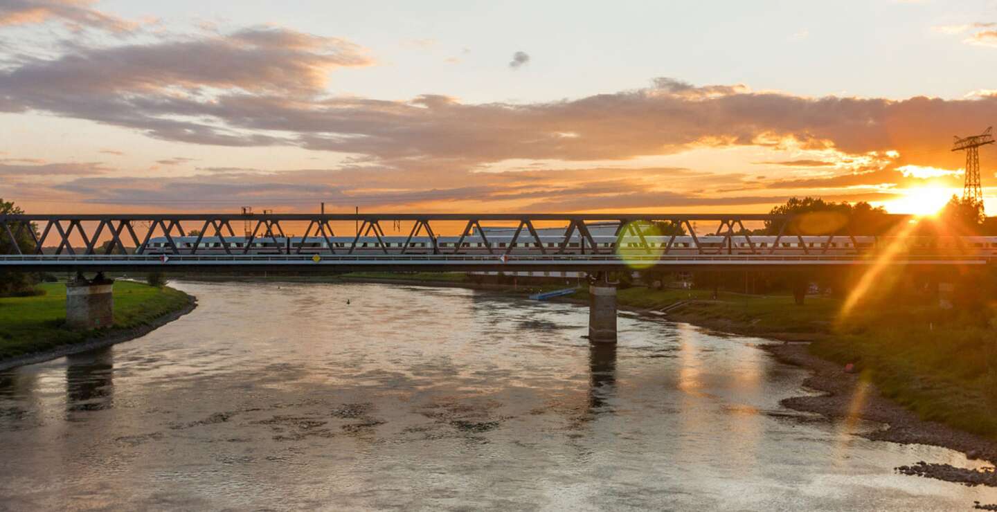 Eine Eisenbahnbrücke überspannt einen Fluss bei Sonnenuntergang, wobei das warme Licht der Sonne auf der Wasseroberfläche reflektiert. | ©  Deutsche Bahn AG