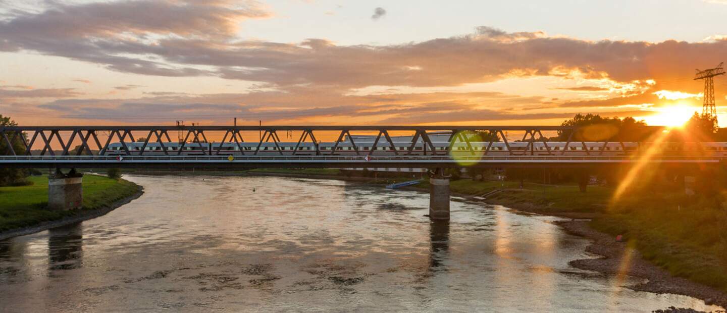 Zug fährt über Brücke im Sonnenuntergang | ©  Deutsche Bahn AG Eine Eisenbahnbrücke überspannt einen Fluss bei Sonnenuntergang, wobei das warme Licht der Sonne auf der Wasseroberfläche reflektiert. | ©  Deutsche Bahn AG