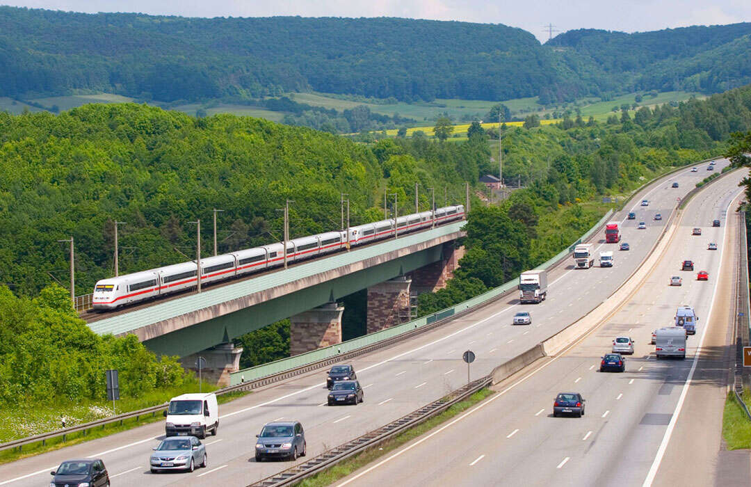 einen modernen Zug, der eine Brücke über eine mehrspurige Autobahn ueberquert, vor einer Kulisse aus gruenen Huegeln und Baeumen. | ©  Deutsche Bahn AG