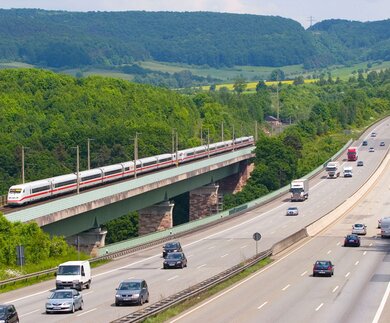 einen modernen Zug, der eine Brücke über eine mehrspurige Autobahn ueberquert, vor einer Kulisse aus gruenen Huegeln und Baeumen. | ©  Deutsche Bahn AG