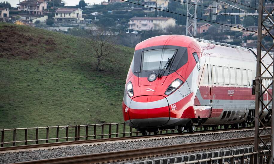Ein rot-weißer Frecciarossa-Zug fährt auf Schienen. Hinter ihm sind Häuser und vor ihm ist eine Landschaft zu sehen. | © Trenitalia