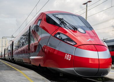 Ein rot-weißer Frecciarossa Zug steht mit geöffneten Türen am Gleis im Bahnhof, Passanten steigen ein. Der Betrachter schaut auf den Zug hinauf, sodass er größer erscheint. | © Trenitalia