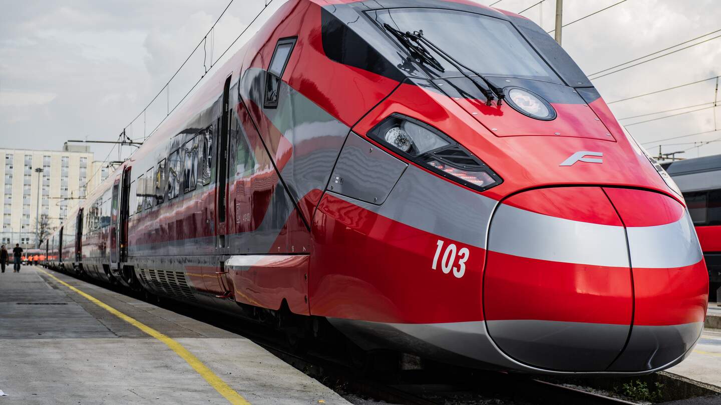 Ein rot-weißer Frecciarossa Zug steht mit geöffneten Türen am Gleis im Bahnhof, Passanten steigen ein. Der Betrachter schaut auf den Zug hinauf, sodass er größer erscheint. | © Trenitalia