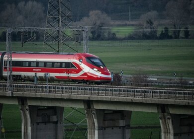 Ein rot-weißer Frecciarossa Zug fährt auf einer Brücke. Neben ihm ist eine Landschaft mit Bäumen zu sehen. | © Trenitalia