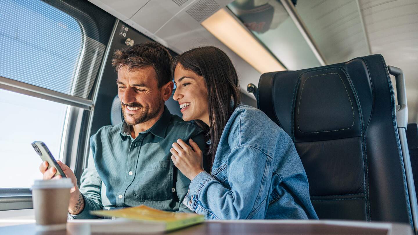 Pärchen im Zug an einem Tisch schauen auf ein Handy | © GettyImages.com/miloradkravic