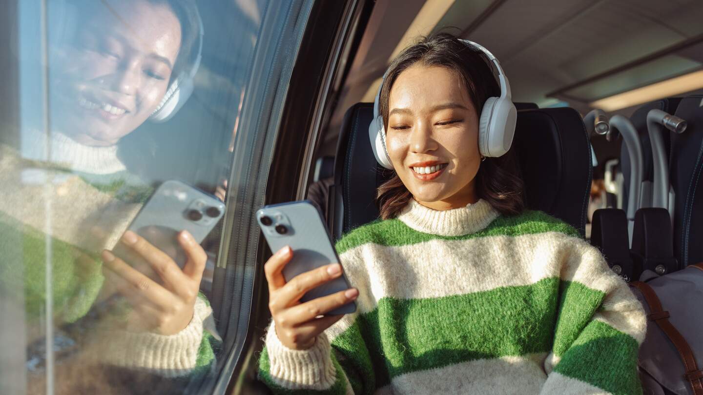 Person sitzt im Zug am Fenster, trägt weiße Kopfhörer und hält ein Smartphone in der Hand, mit Spiegelung im Glas und Tageslicht im Hintergrund. | © Gettyimages.com/	Milko