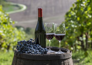Fass auf dem eine Flasche Rotwein und zwei gefuellte Rotwein Glaeser stehen, daneben liegen Trauben und im Hintergrund sind Weinberge zu sehen | © Gettyimages.com/Alberto Gagliardi