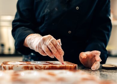 Nahaufnahme der Haende eines Konditors, der weisse Pralinen in einer handwerklichen Werkstatt schmueckt, Bruessel | © Gettyimages.com/Carlo Prearo