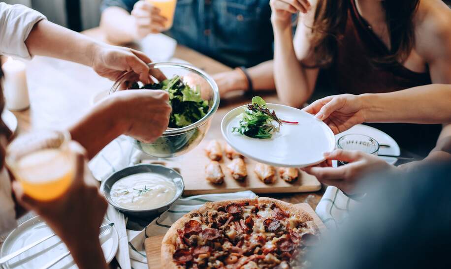 Nahaufnahme einer jungen Gruppe fröhlicher Freunde, die Spaß daran haben, zusammen zu genießen, Essen über den Esstisch auf der Party zu verteilen und zu teilen | © © Gettyimages.com/AsiaVision