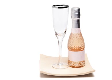 Nahaufnahme einer kleinen Flasche Champagner neben einem edlen Glas mit silbernem Rand auf einer leicht geschwungenen Kiefernholzplatte auf weissem Hintergrund | © Gettyimages.com/PictureSyndicate