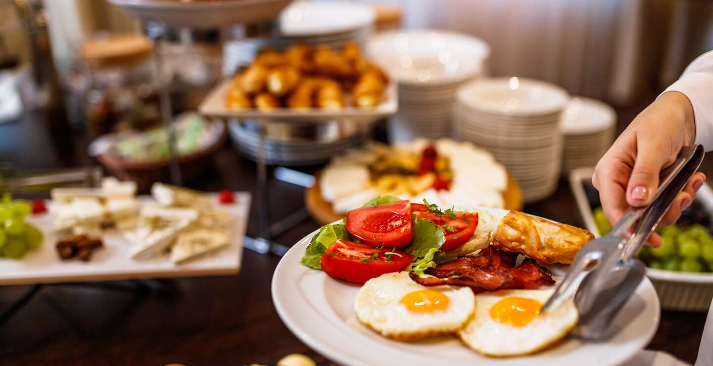 Frühstücksteller mit zwei Spiegeleiern und dem Restaurantbuffet im Hintergrund | © gettyimages.com/MilanEXPO