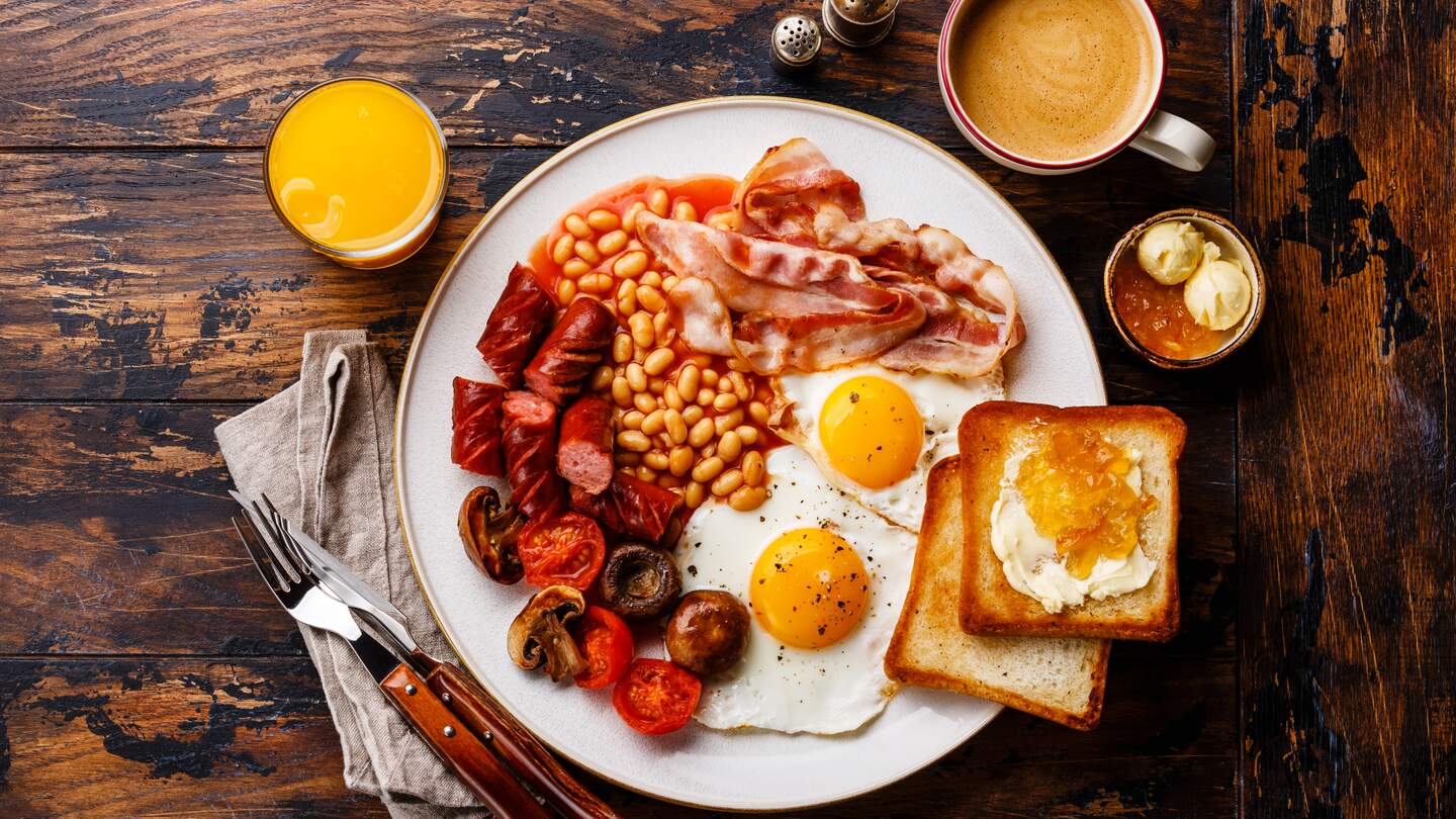 Englisches Frühstück mit Spiegeleiern, Würstchen, Speck, Bohnen, Toast und Kaffee auf Holzhintergrund. | © GettyImages.com/Lisovskaya