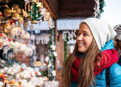Froehliche junge Frau betrachtet bunte Weihnachtsschmuck-Auslagen an einem Holzhuettestand auf dem Weihnachtsmarkt bei schoenem Winterwetter | © Gettyimages.com/Zbynek Pospisil