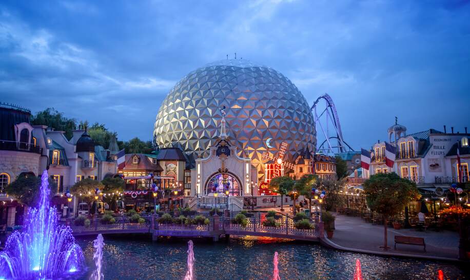 Beleuchtete Glaskuppel inmitten einer Parklandschaft mit farbigen Springbrunnen und Häuserfassaden, die bei Abendstimmung eine magische Freizeitpark-Atmosphäre schaffen. | © Europa-Park