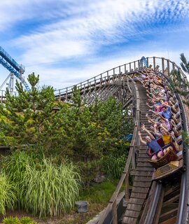 Holzachterbahn rauscht mit voller Geschwindigkeit durch eine steile Kurve, umgeben von grüner Landschaft, während Fahrgäste den intensiven Nervenkitzel genießen. | © Europa Park