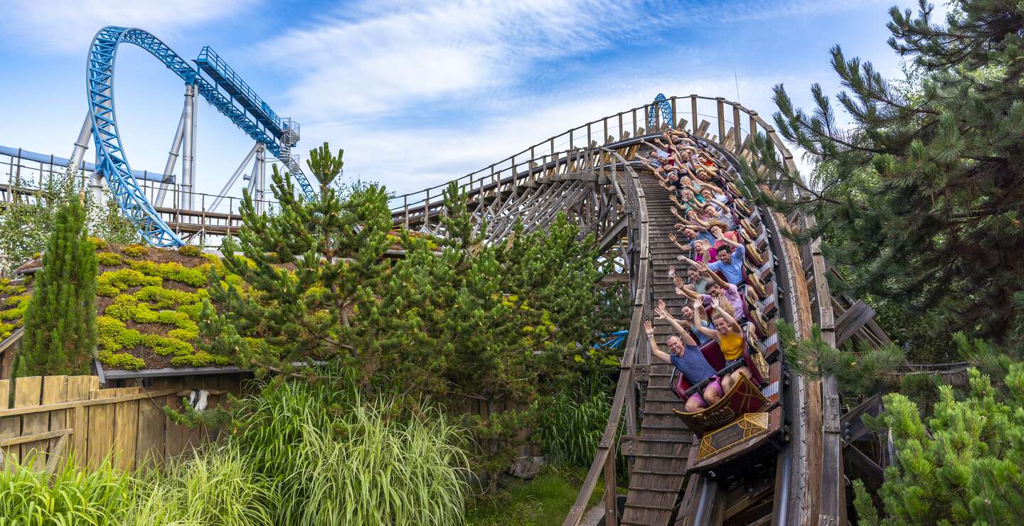 Holzachterbahn rauscht mit voller Geschwindigkeit durch eine steile Kurve, umgeben von grüner Landschaft, während Fahrgäste den intensiven Nervenkitzel genießen. | © Europa Park