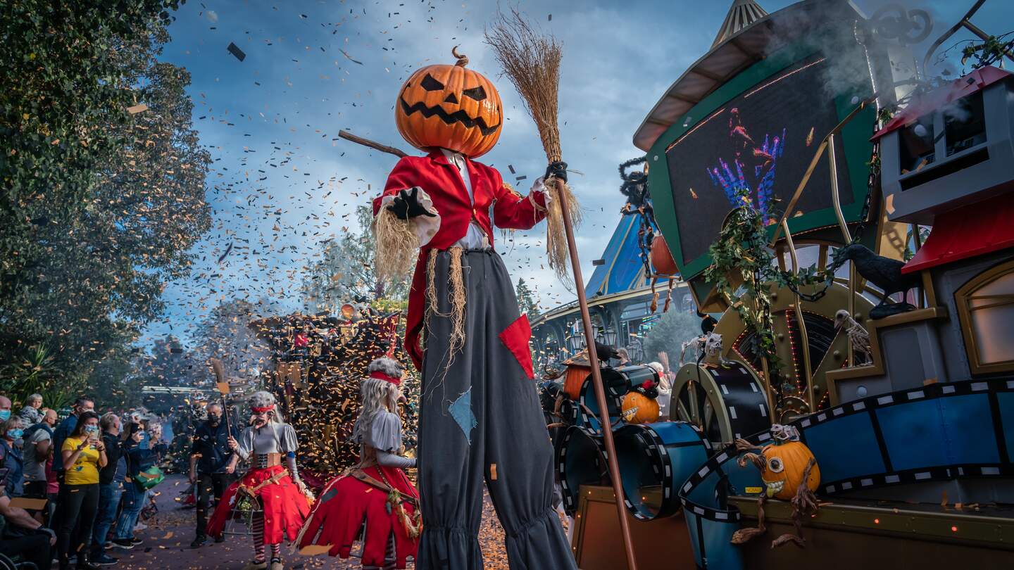 Übergroße Kürbisfigur auf Stelzen mit Besen während einer Halloween-Parade im Europa-Park, umgeben von Zuschauern und dekoriertem Festwagen. | © Europa-Park