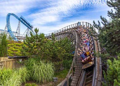 Holzachterbahn rauscht mit voller Geschwindigkeit durch eine steile Kurve, umgeben von grüner Landschaft, während Fahrgäste den intensiven Nervenkitzel genießen. | © Europa Park