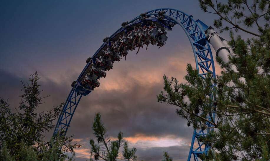 Die Bluefire-Achterbahn fährt bei Sonnenuntergang durch einen hohen Looping, umgeben von Bäumen und dramatischem Himmel, und erzeugt ein actionreiches Fahrerlebnis. | © Gettyimages.com/wirestock