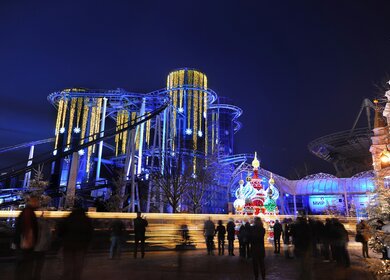 Beleuchtete Achterbahn im nächtlichen Freizeitpark, umgeben von winterlicher Dekoration und Besuchern, die die stimmungsvolle Atmosphäre im funkelnden Lichterglanz genießen. | © Europa-Park 