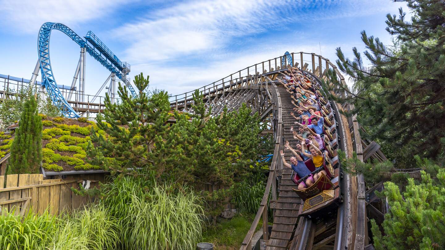 Holzachterbahn rauscht mit voller Geschwindigkeit durch eine steile Kurve, umgeben von grüner Landschaft, während Fahrgäste den intensiven Nervenkitzel genießen. | © Europa Park