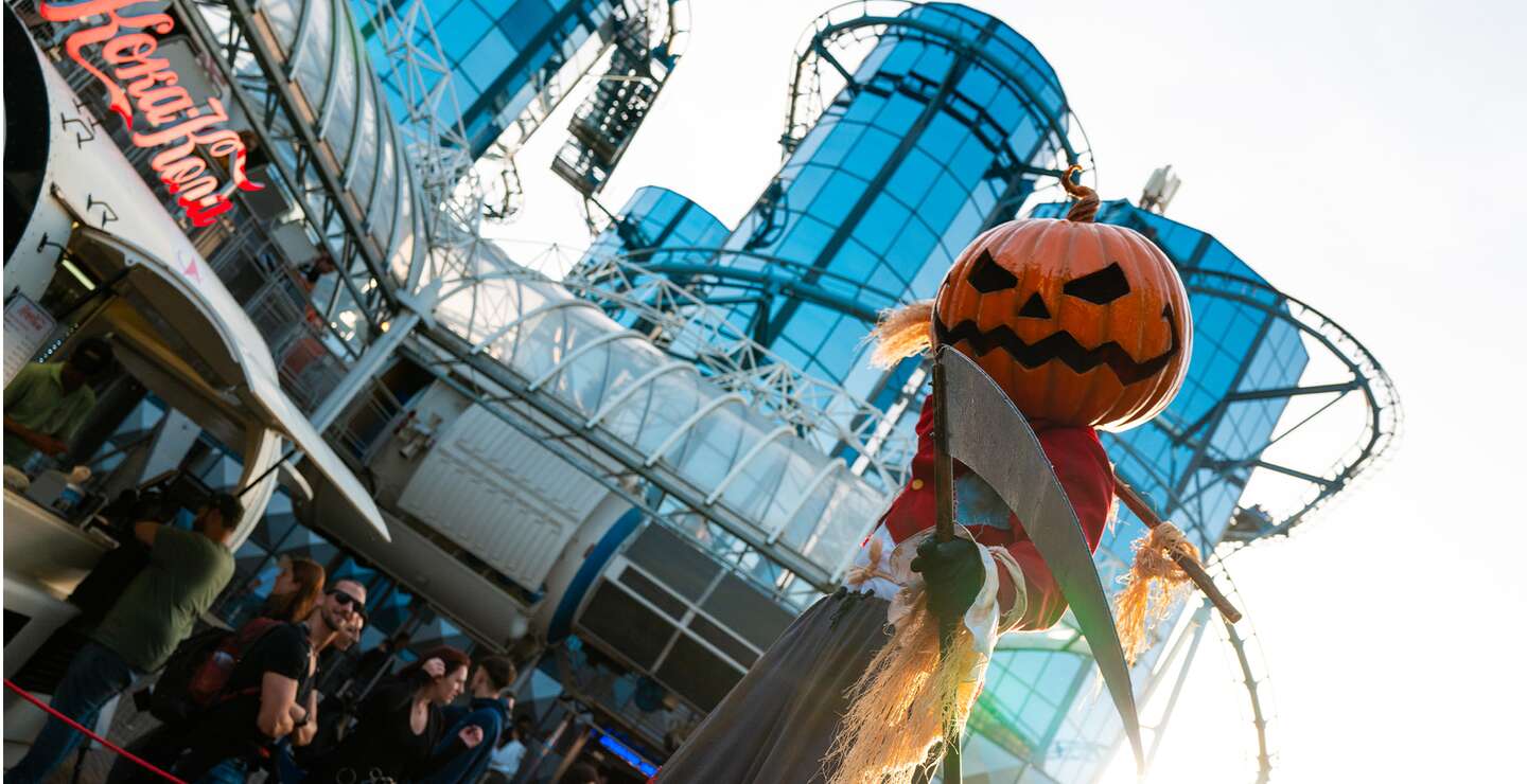 Halloween-Dekoration im Europa-Park mit einer großen Kürbisfigur in Vogelscheuchengestalt vor einem modernen Gebäude mit Glasfassade, die Achterbahn Euro-Mir im Hintergrund. | © Europa-Park