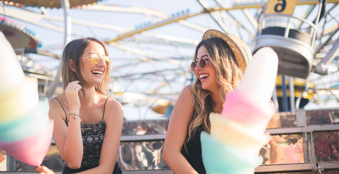 Spaß im Freizeitpark: Freundinnen genießen Zuckerwatte vor Achterbahnkulisse | © gettyimages.com/DusanManic